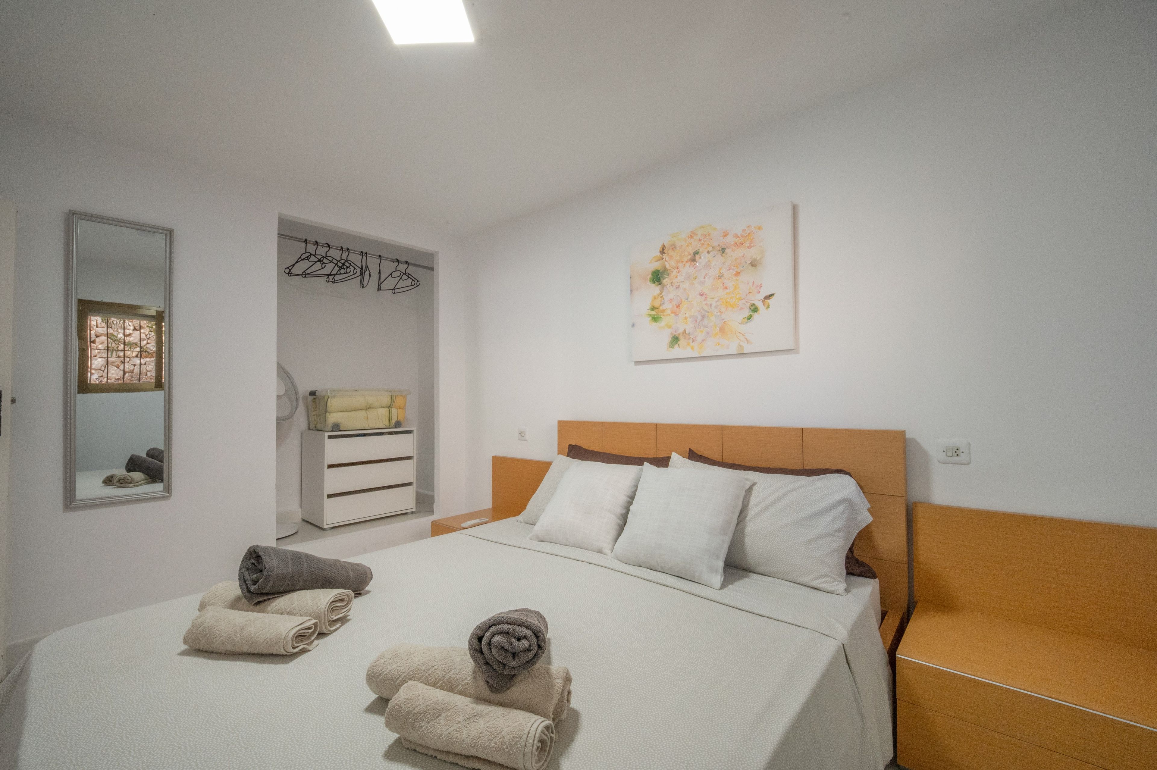 Habitación de hotel con cama, cabecera de madera, mesita de noche, espejo, armario y un cuadro en la pared.