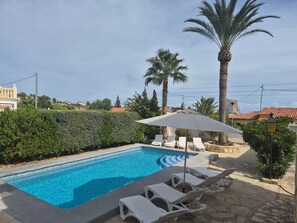 Ein Swimmingpool mit Liegestühlen und Sonnenschirm, umgeben von Palmen und einer Hecke.