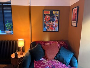 Ein Zimmer mit einer Couch, einer Lampe, einem gerahmten Poster und einem Blumentopf.