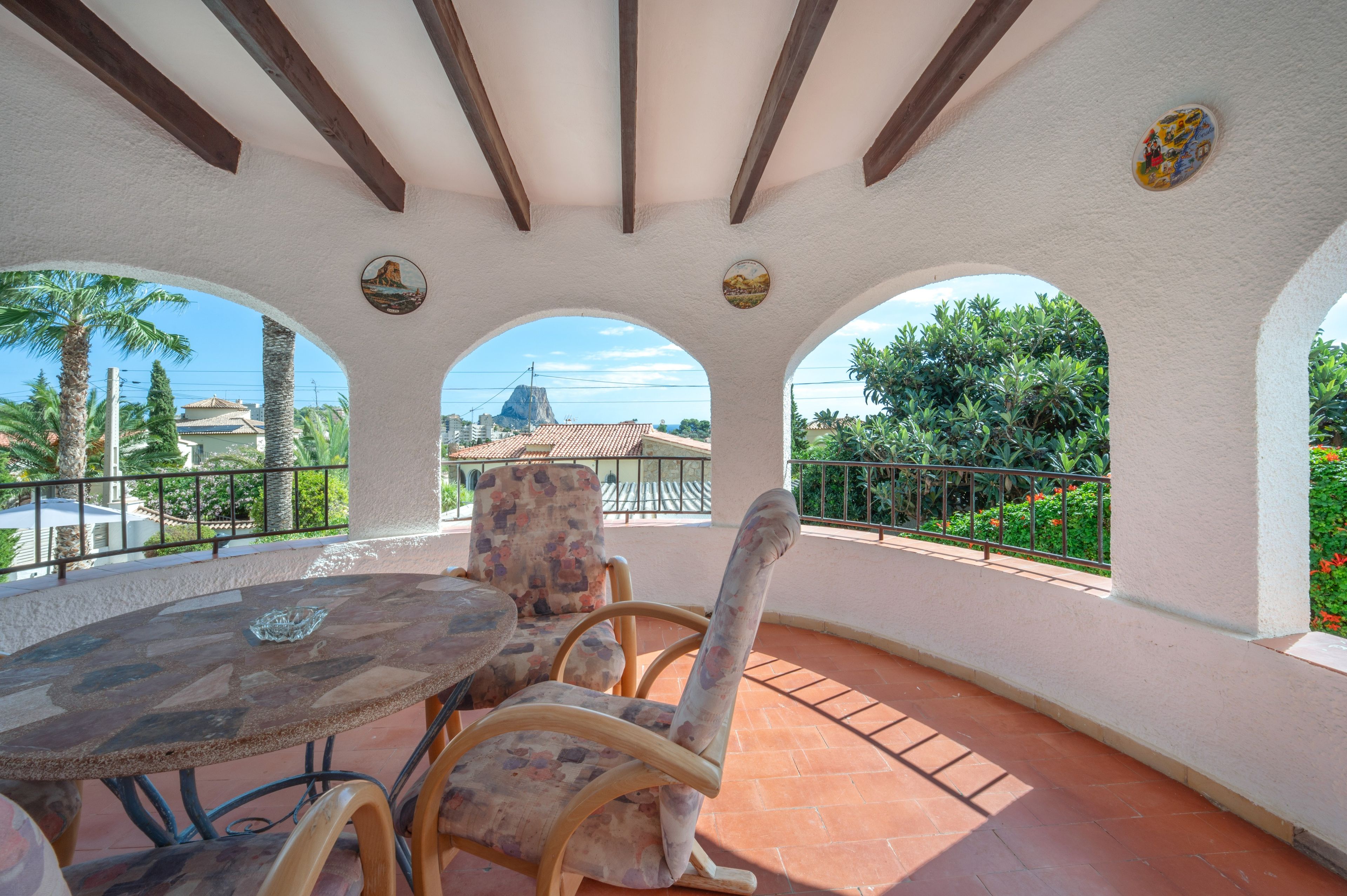 A covered patio with a round table and chairs, a stone pillar, and a view of a mountain and buildings.