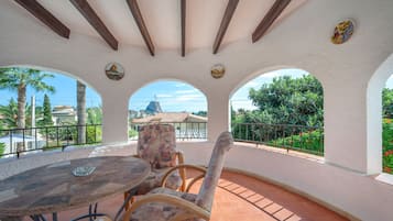 A covered patio with a round table and chairs, a stone pillar, and a view of a mountain and buildings.