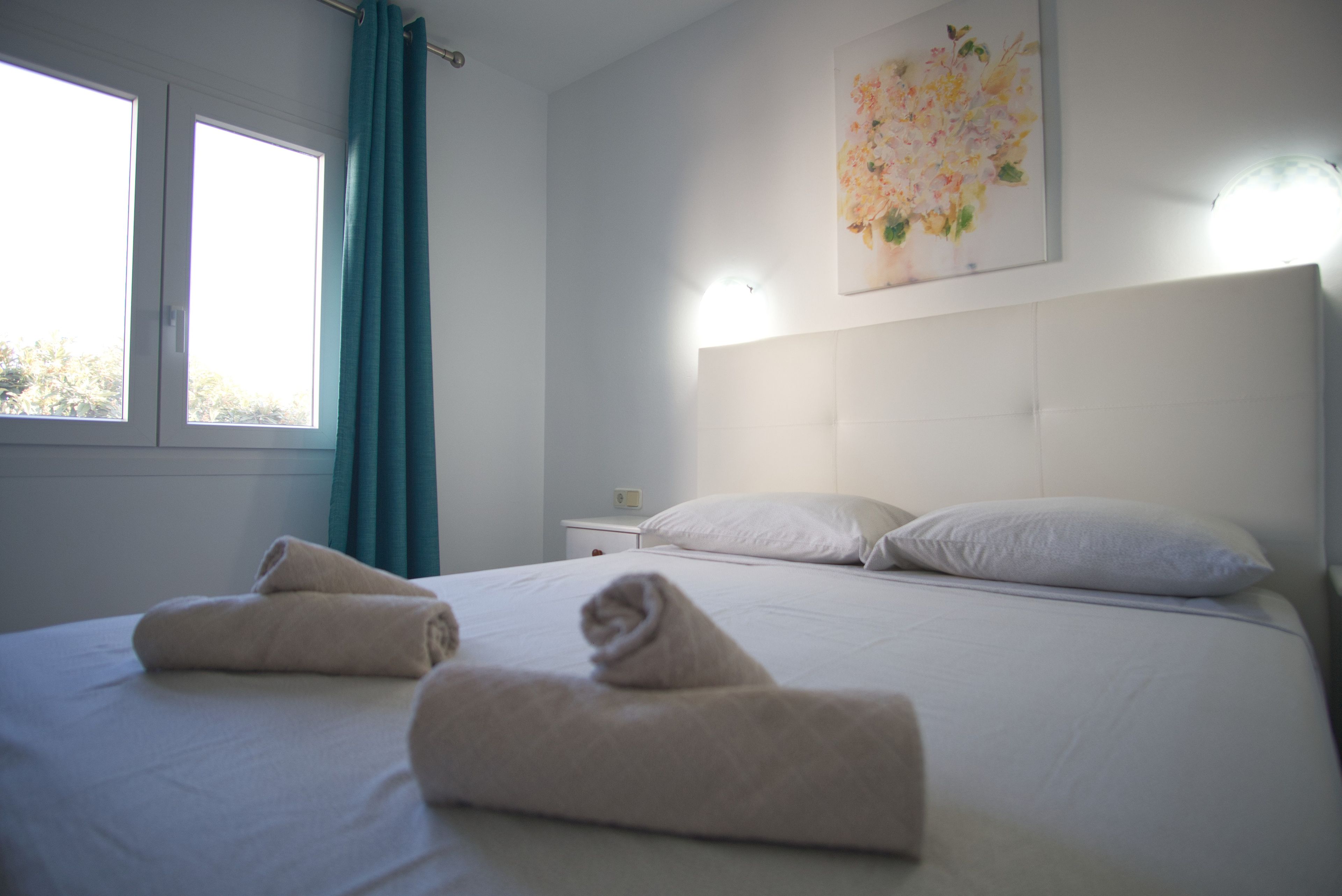 Een hotelkamer met een bed, twee kussens, een nachtkastje, een aan de muur hangend schilderij en een raam met gordijnen.