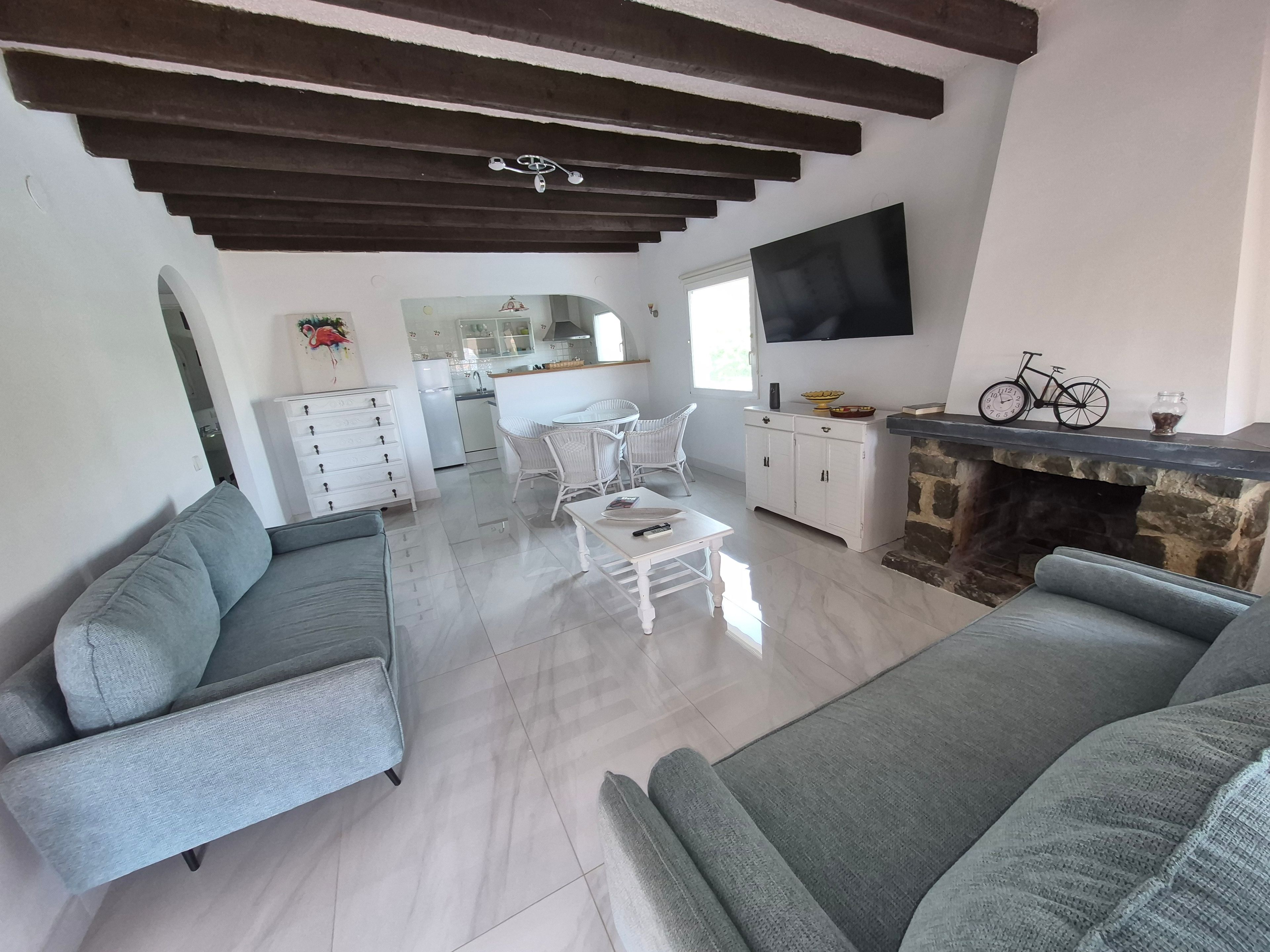A modern living room with grey sofas, a dining area with white chairs, a kitchen with a stone fireplace, and a flat-screen TV mounted on the wall.