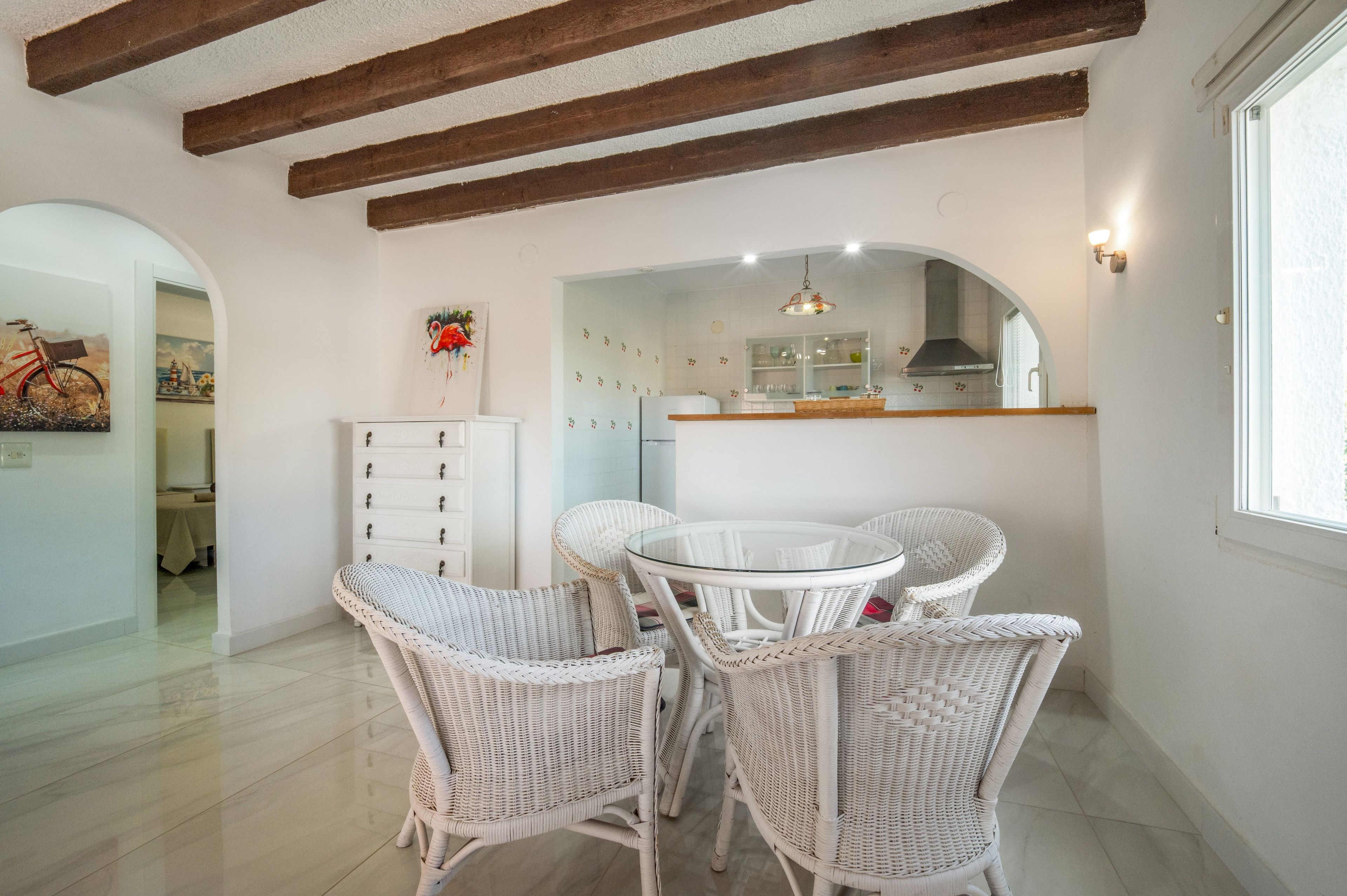 A dining area with wicker chairs, a glass-top table, and a kitchen in the background.