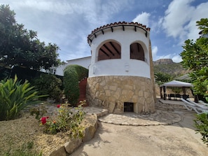 Edifici amb base de pedra i parets superiors blanques, zona d'esbarjo coberta a l'aire lliure i jardí amb plantes i flors.