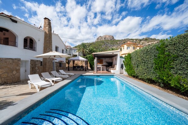 A swimming pool with lounge chairs, a stone building, and a mountain in the background.