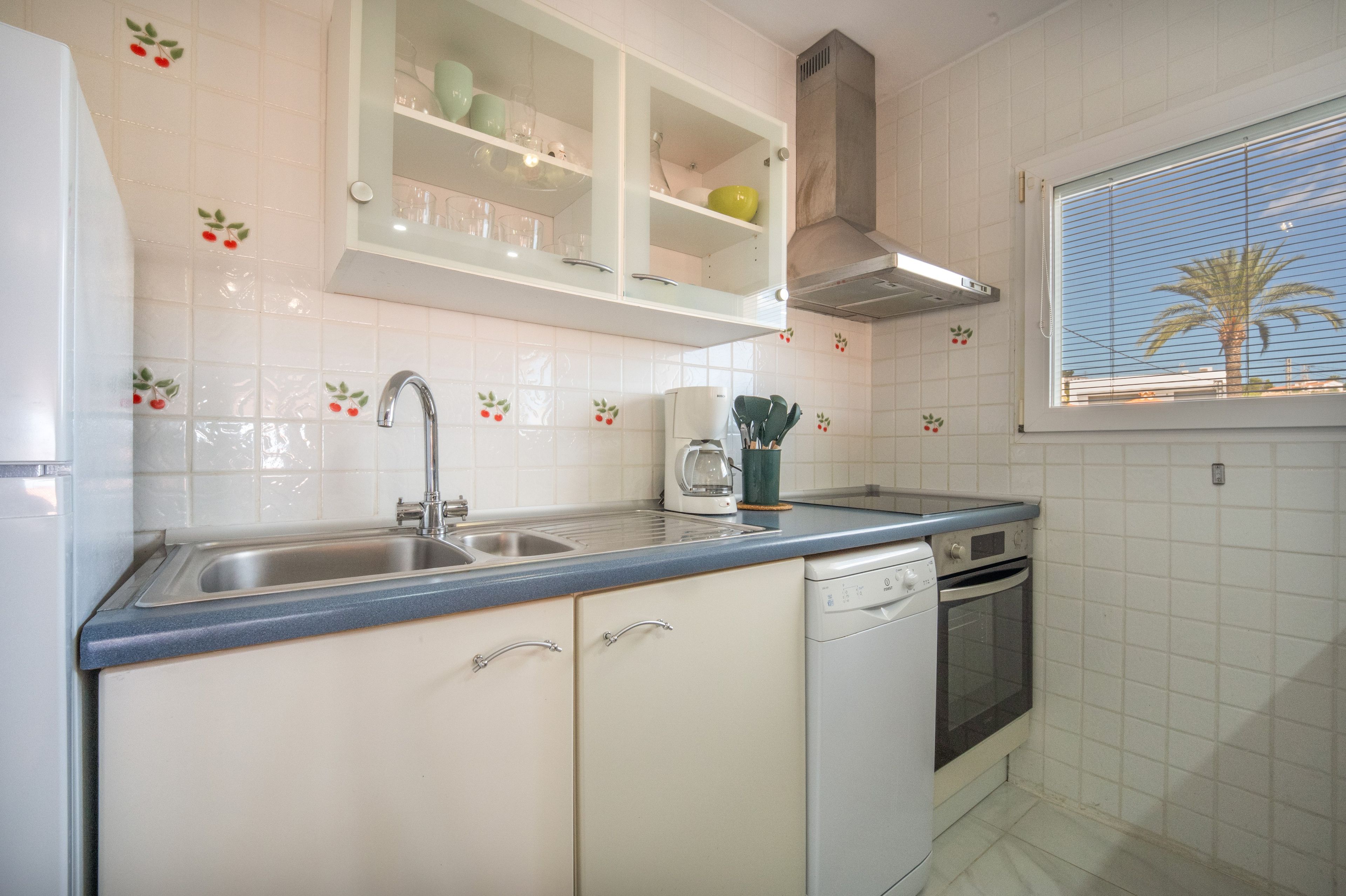 Una cucina con mobili bianchi, un lavello doppio, un fornello e una finestra con vista su palme.