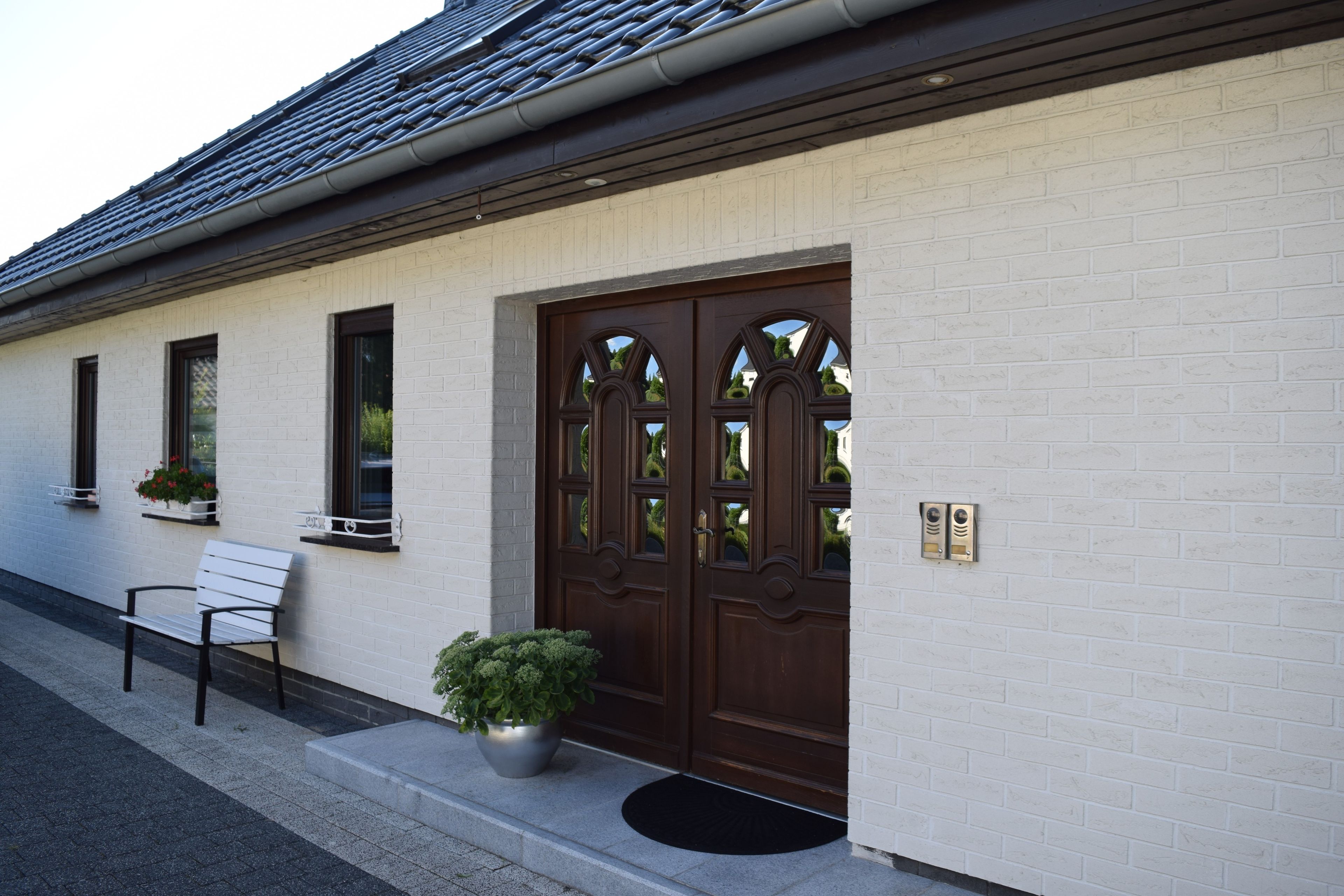Ein Gebäude mit weiß verputzter Fassade, einer braunen Tür mit Glasfeldern, einer Bank und einem Blumentopf.