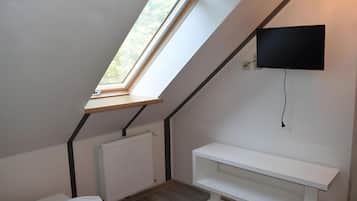 Ein Zimmer mit schrägem Dach, einem Fenster, einer Heizung, einem an der Wand befestigten Fernseher und einer weißen Bank.