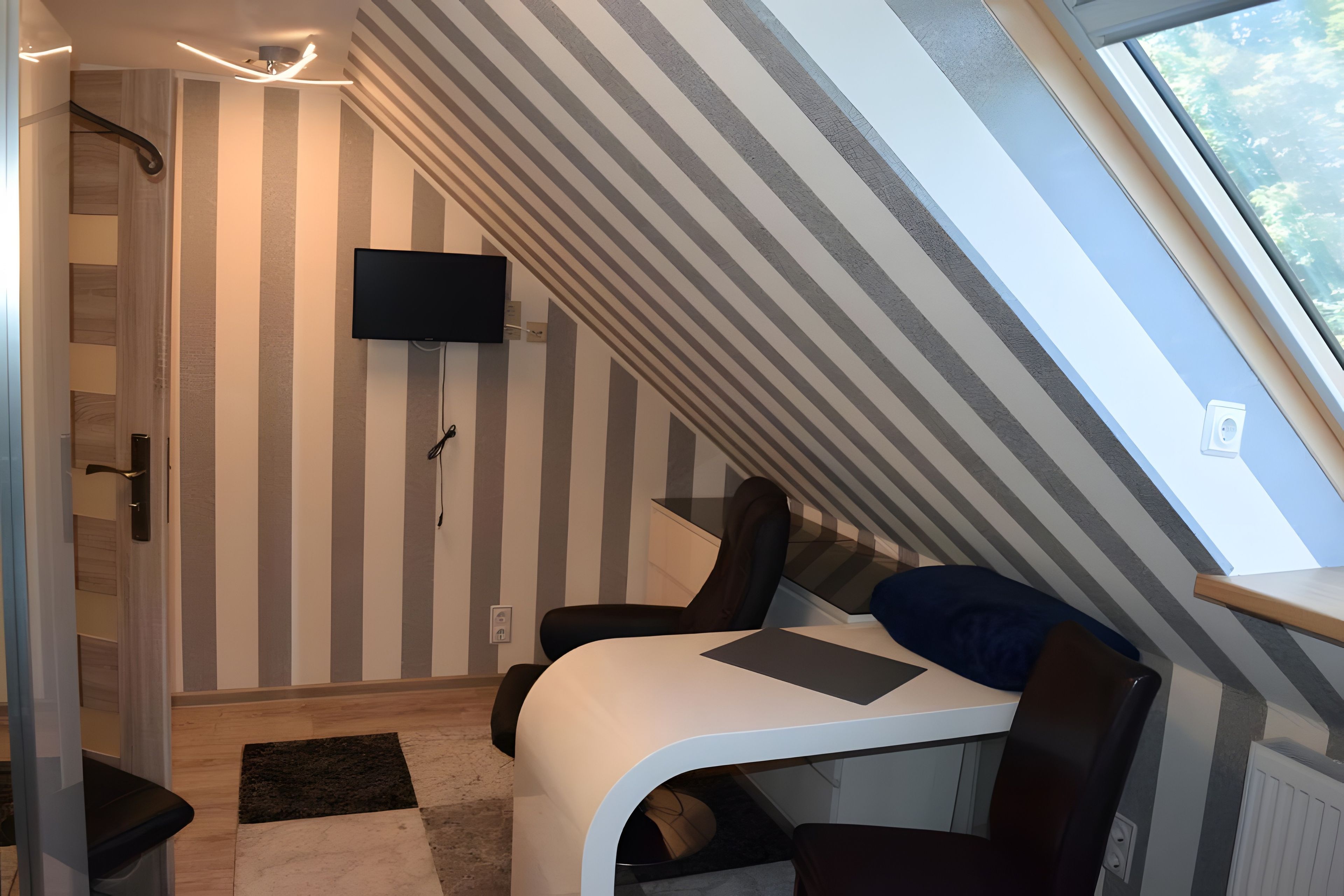 Une pièce dotée d’un plafond en pente, d’un téléviseur à écran plat fixé au mur, d’un bureau avec une chaise et d’une fenêtre donnant sur l’extérieur.