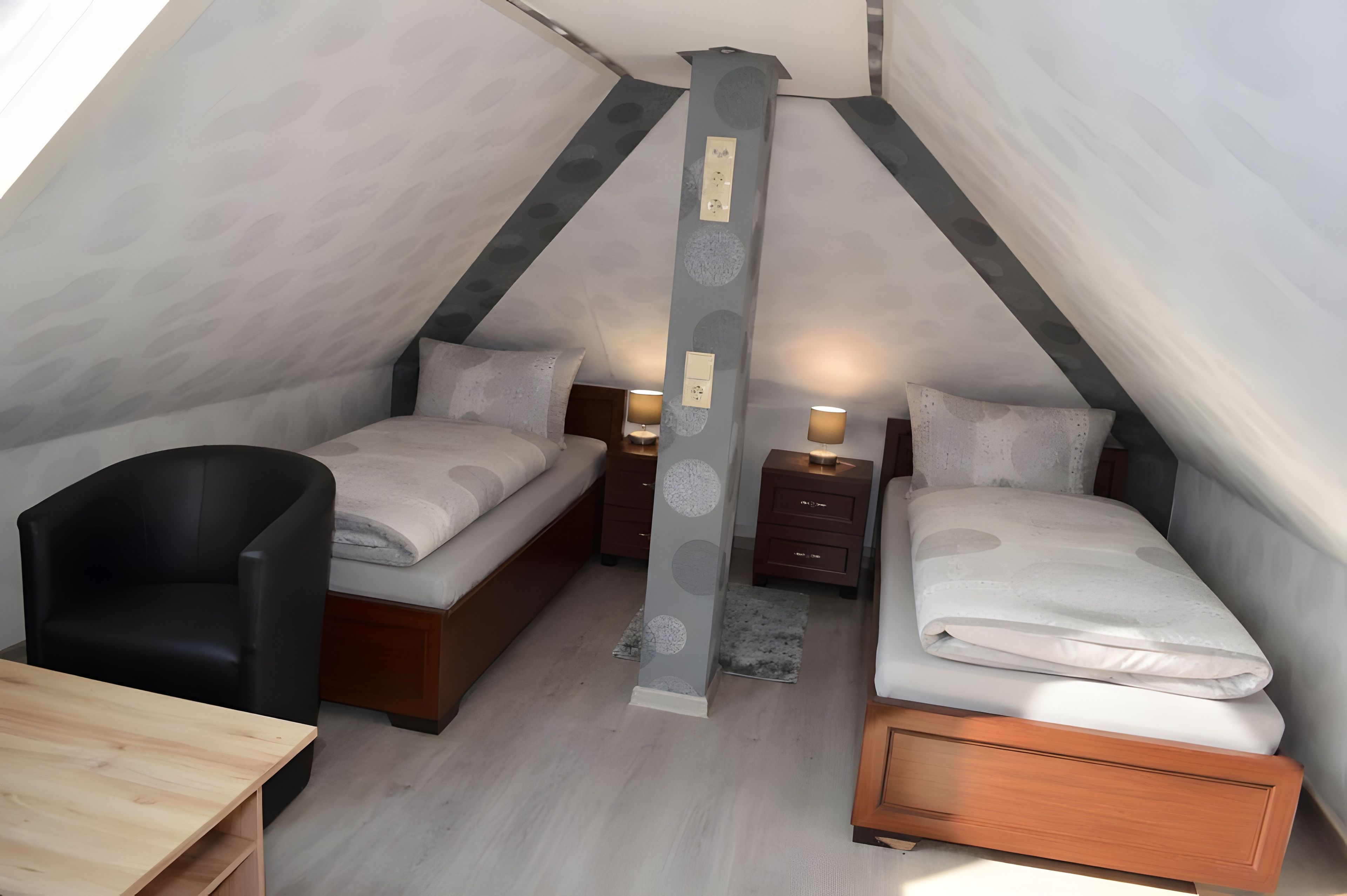 Une petite chambre mansardée de forme triangulaire, avec deux lits, un bureau et une chaise.