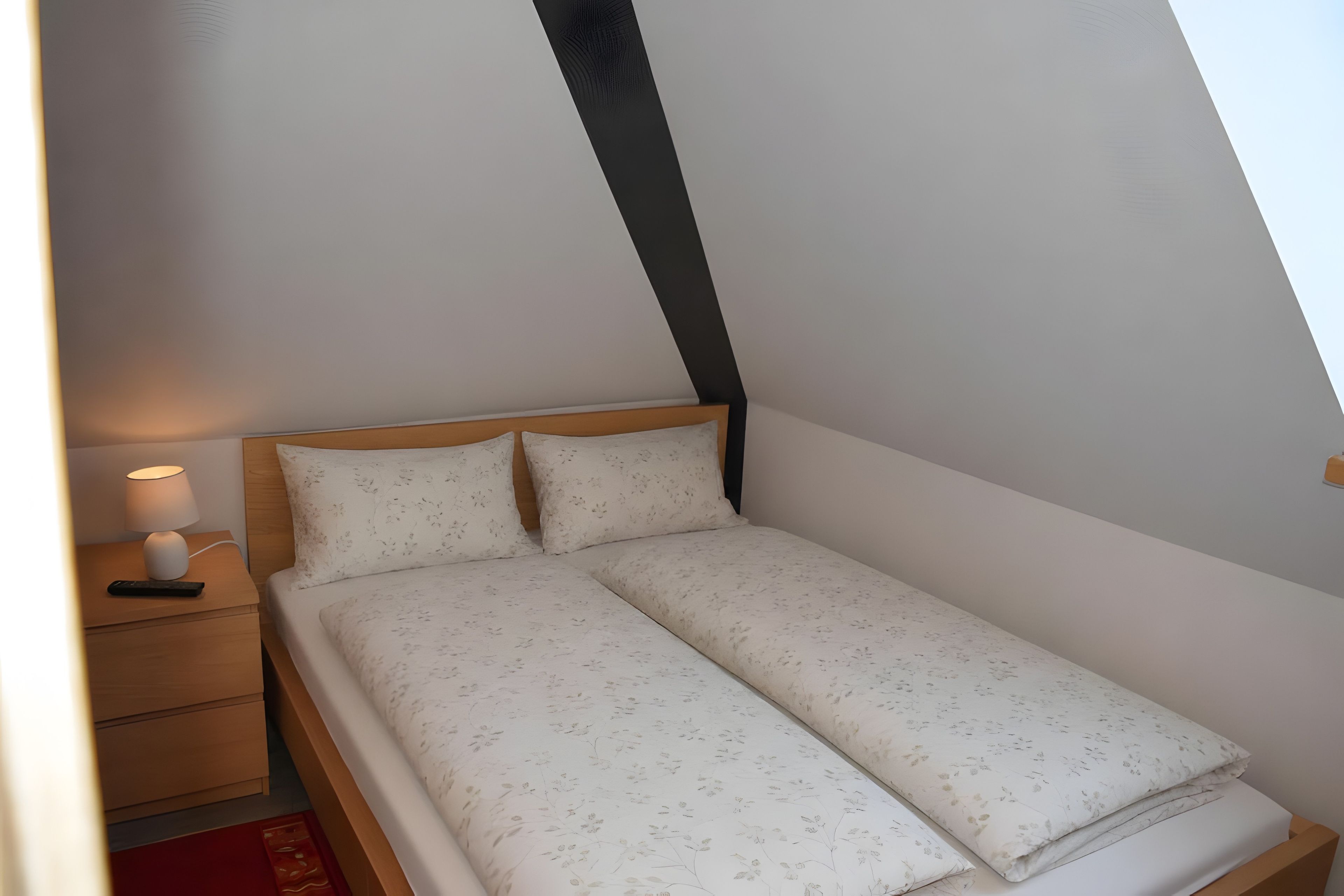 Una habitación pequeña con cama individual, techo inclinado, mesita de noche con lámpara y control remoto.