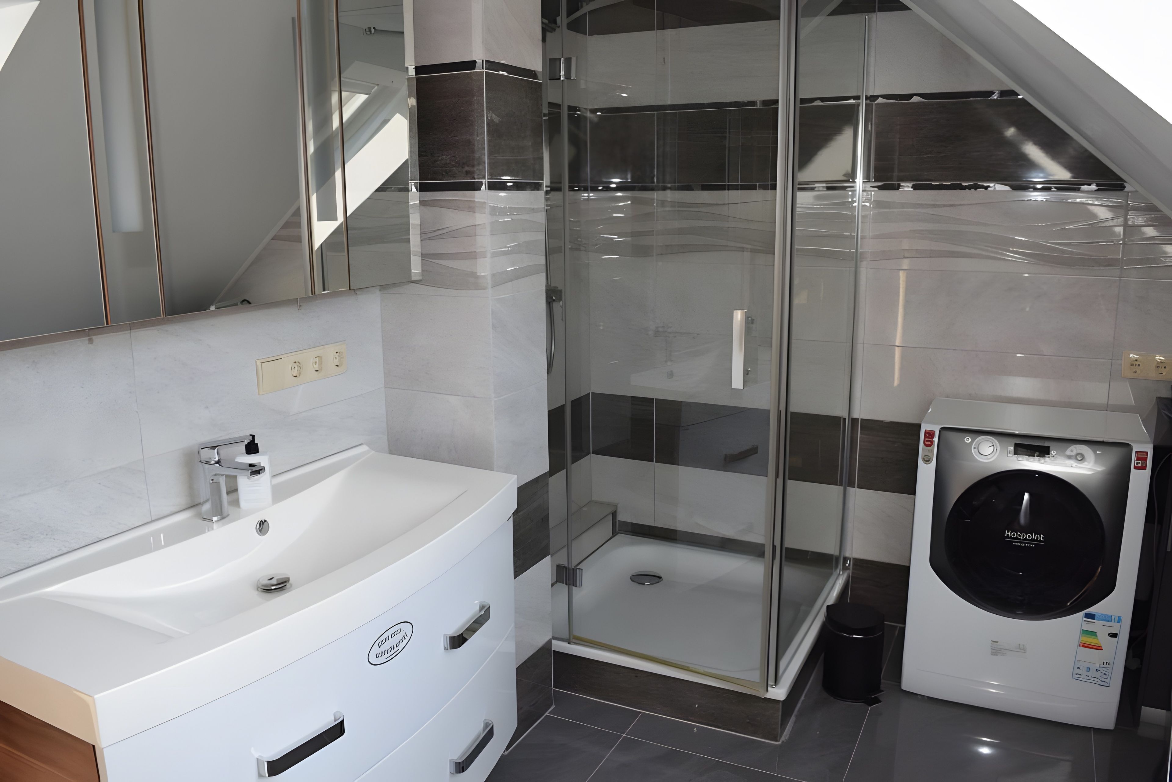 Ein modernes Badezimmer mit einem weißen Waschbecken, einer Duschkabine aus Glas und einer integrierten Waschmaschine.