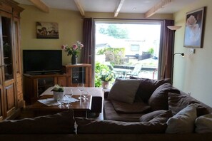 Ein Wohnzimmer mit einer Couch, einem Fernseher, einem Esstisch mit Weingläsern und Blick durch eine Glastür.