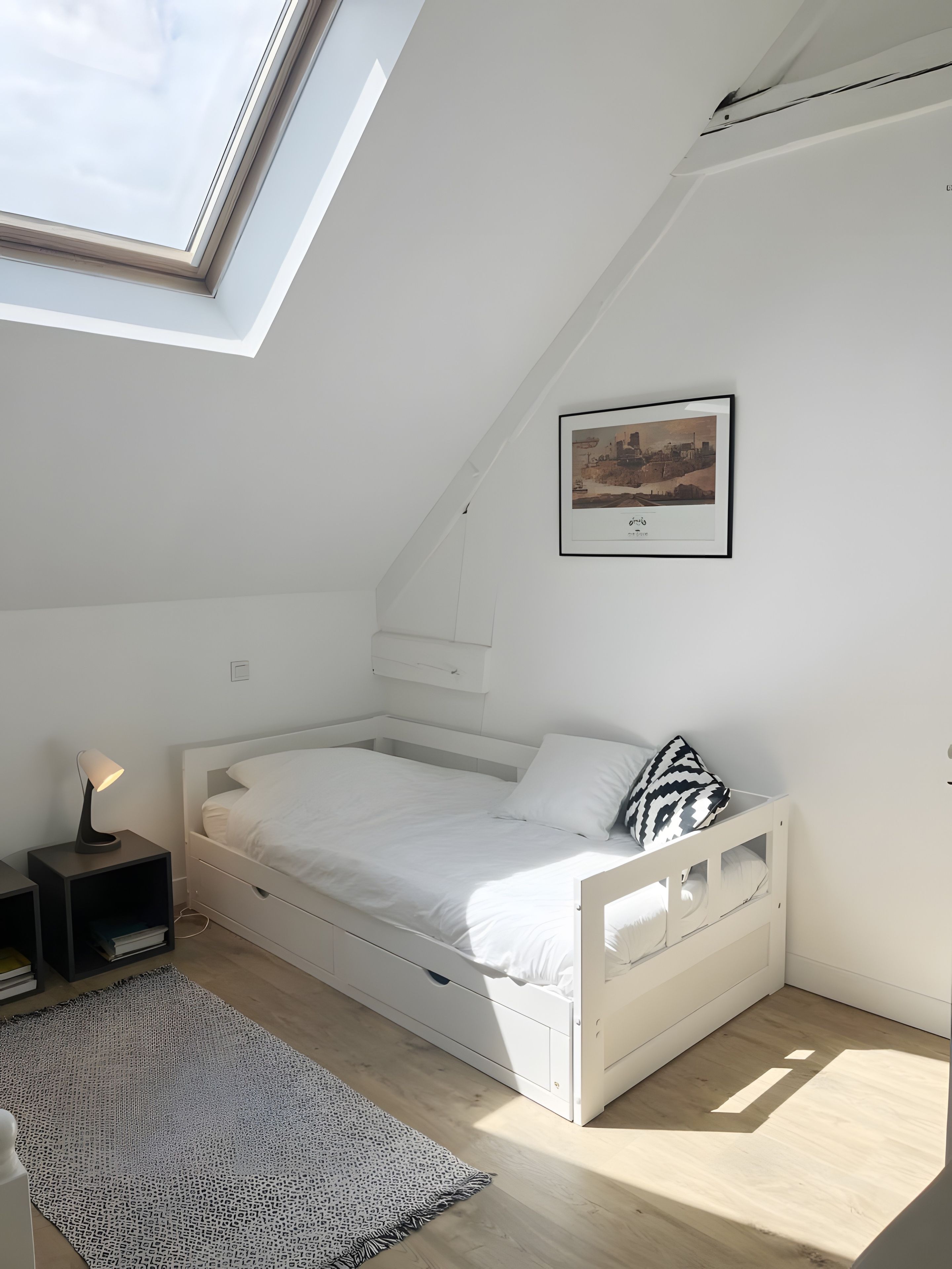 Een slaapkamer met dakraam, een bed, een nachtkastje met lamp, een kamerkleed met patroon, en een ingelijst beeld aan de muur.