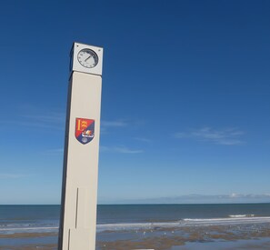 A tall clock tower with a clock face and a crest on its side, overlooking a beach with waves and a clear sky. - Bed and Breakfast 'L'échappée Belle - Chambres' with Wi-Fi (Luc-sur-Mer)