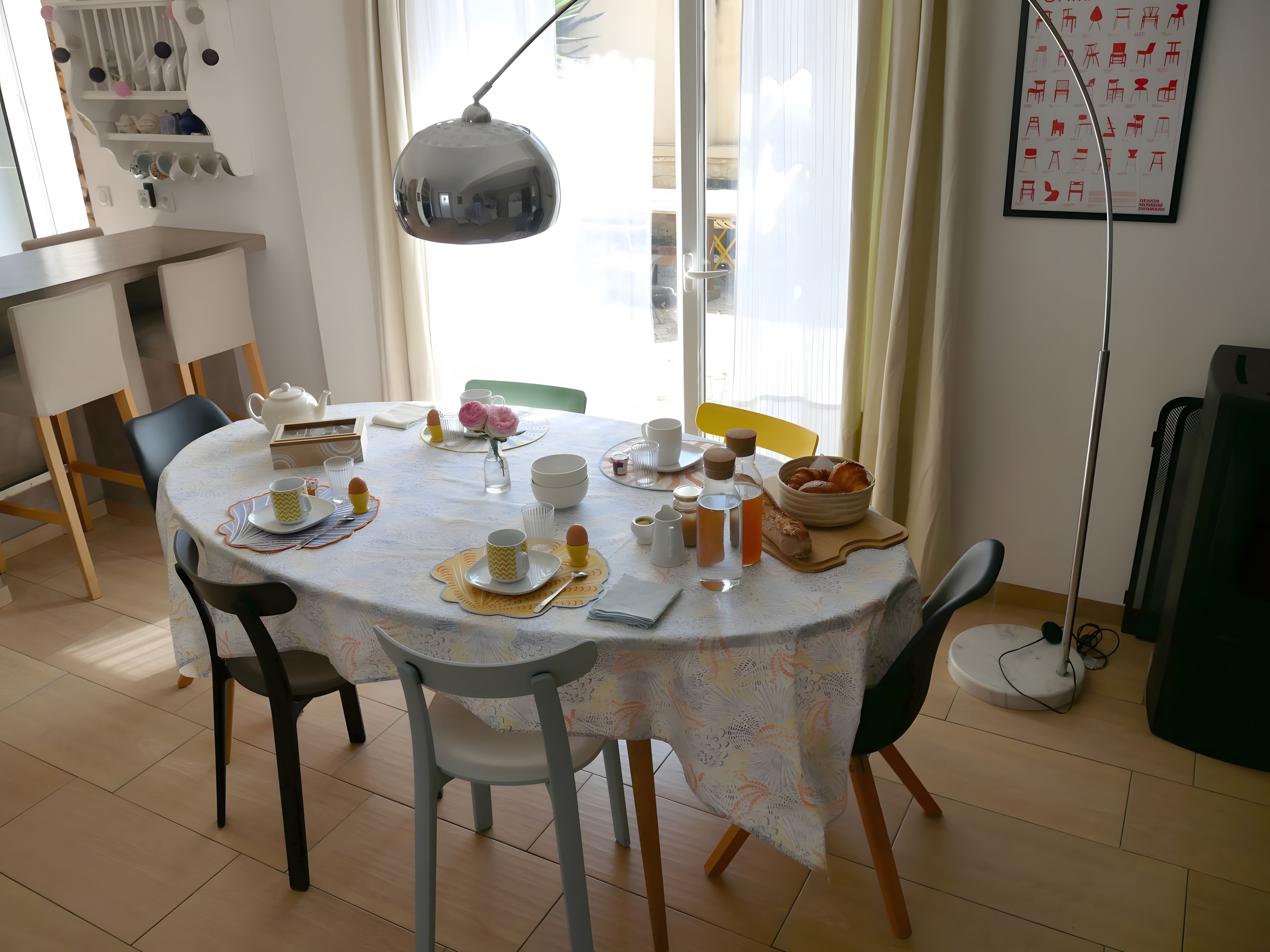 A dining area with a round table set for four, chairs, a lamp, and a wall-mounted shelf with cups and a teapot.