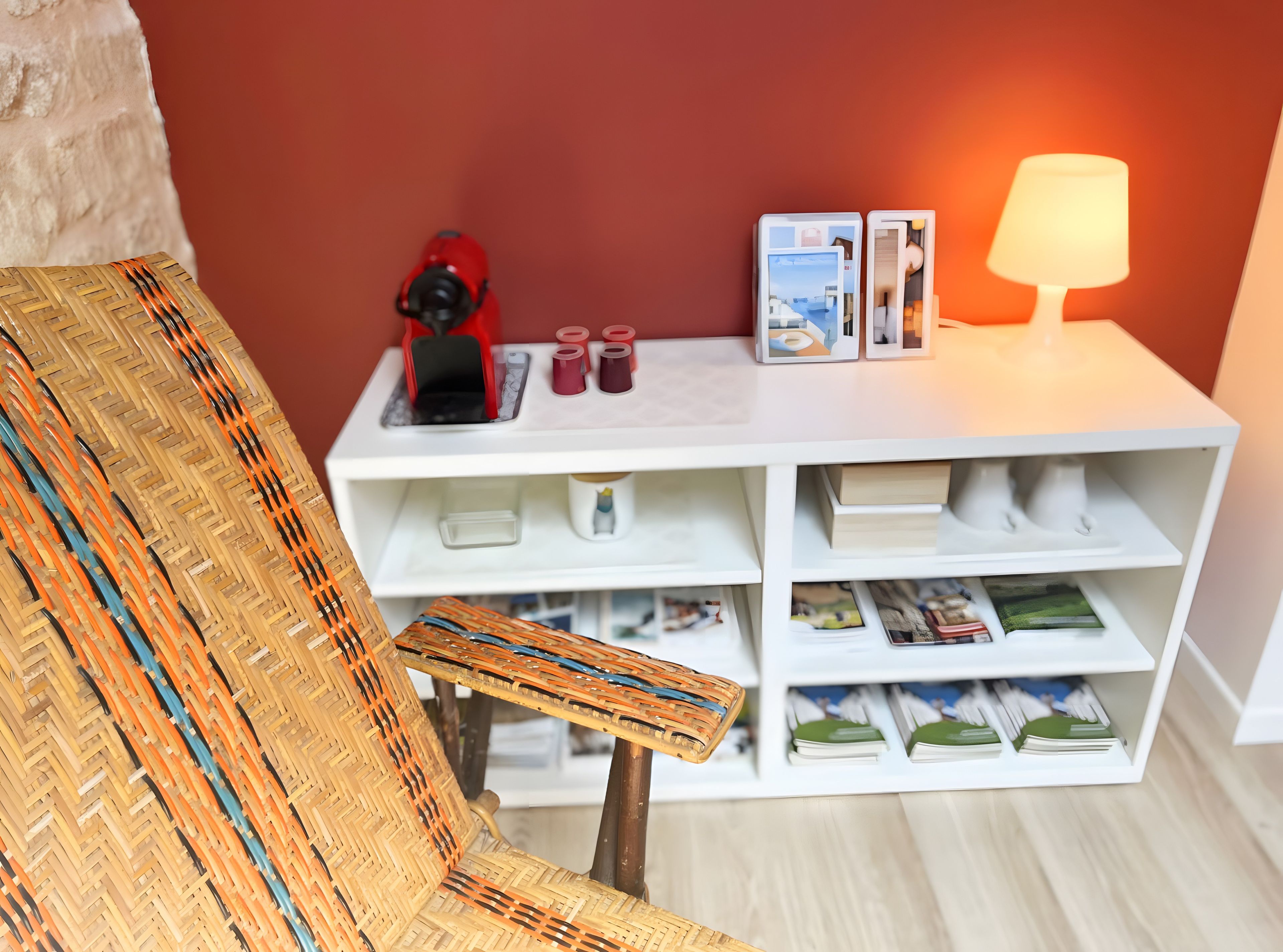 A room with a wicker chair, a white shelf with books and decorative items, and a lamp.