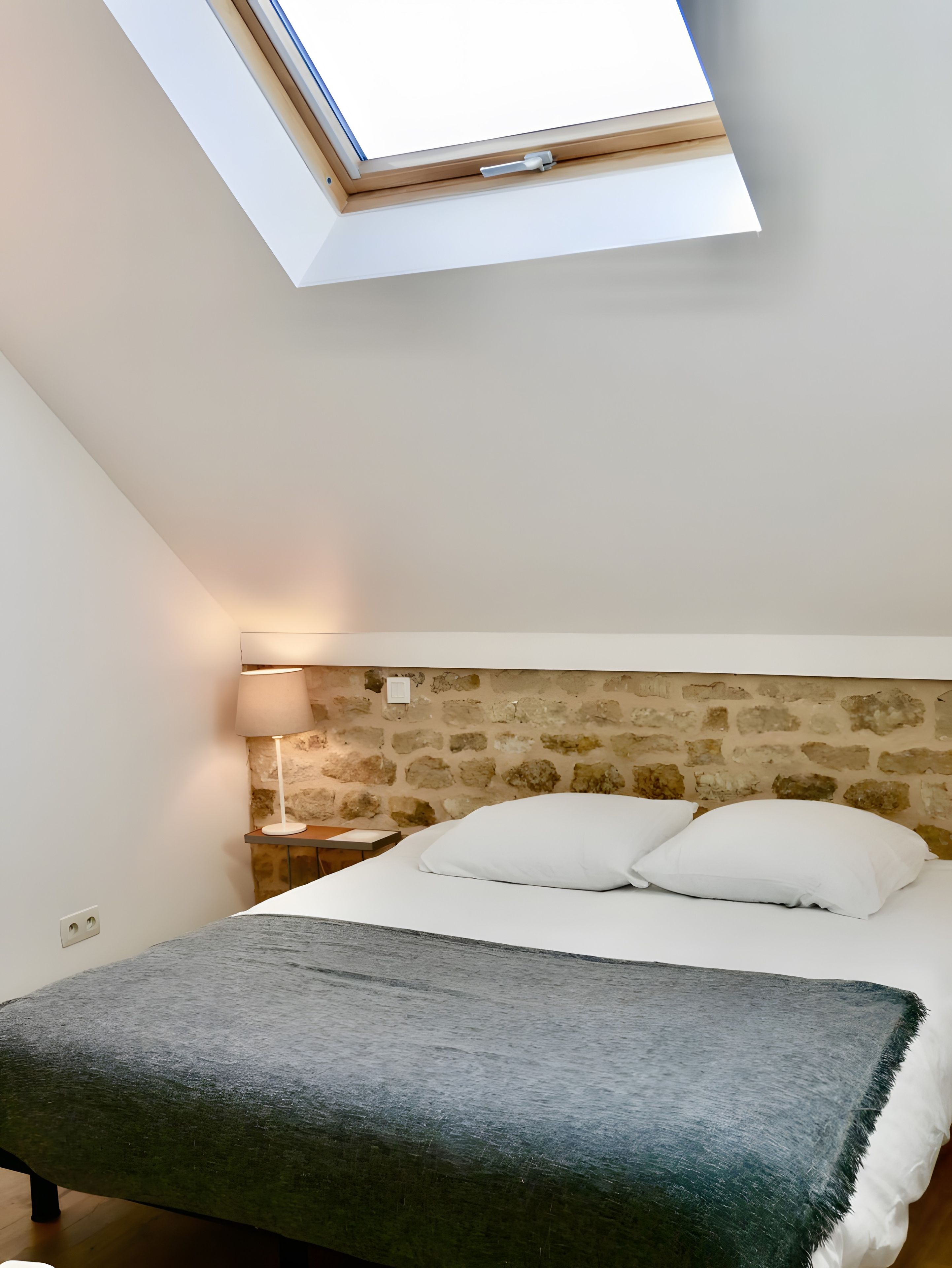 A bedroom with a skylight, stone wall, bed with pillows, bedside lamp, and a bedside table.