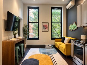 A modern kitchen with a yellow sofa, a dining area with stools, and large windows with a view of trees. - Stylish Downtown 1BR | Walkable to NFL (Pittsburgh)