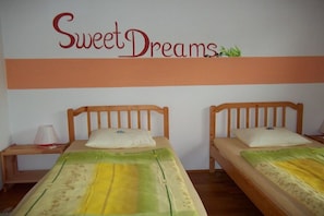Zwei Einzelbetten mit Holzböden und weißer Bettwäsche, ein Nachttisch mit Lampe sowie eine Wand mit der Aufschrift 'Sweet Dreams' über einem horizontalen, orangefarbenen Streifen.