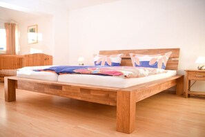 A wooden bed with a colorful blanket and pillows, a wooden nightstand with a lamp, and a wooden bench in a bright room with a wooden floor. - Holiday apartment with 1 bedroom (Titisee-Neustadt)