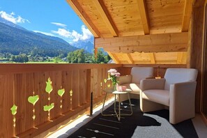 A wooden balcony with a view of mountains, a round table with a vase of flowers, and two chairs. - Holiday apartment with 1 bedroom (Schönau am Königssee)