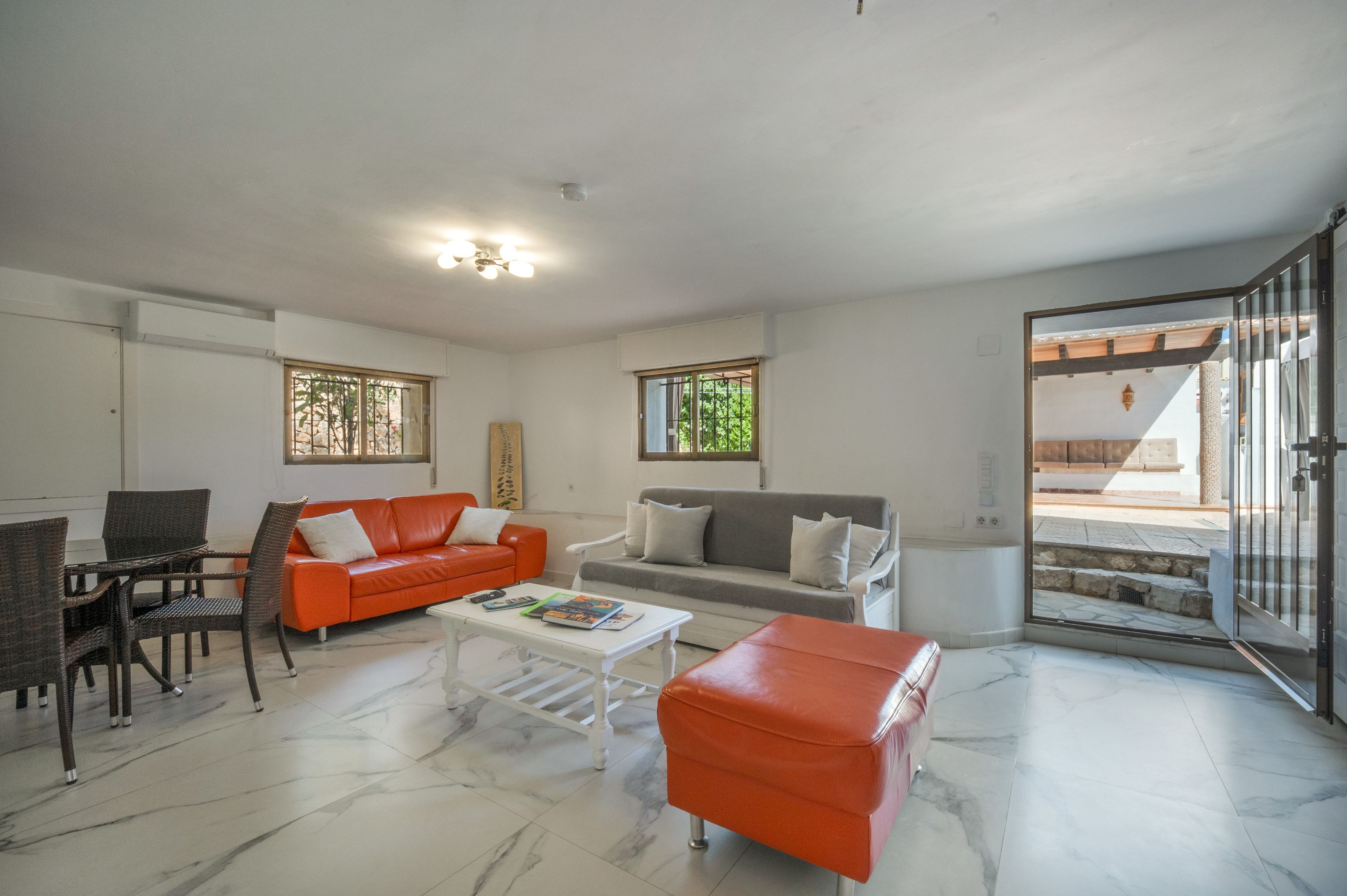 A living room with a sofa, armchairs, a coffee table, and a red ottoman. There are windows with blinds, a ceiling light, and a door leading outside.
