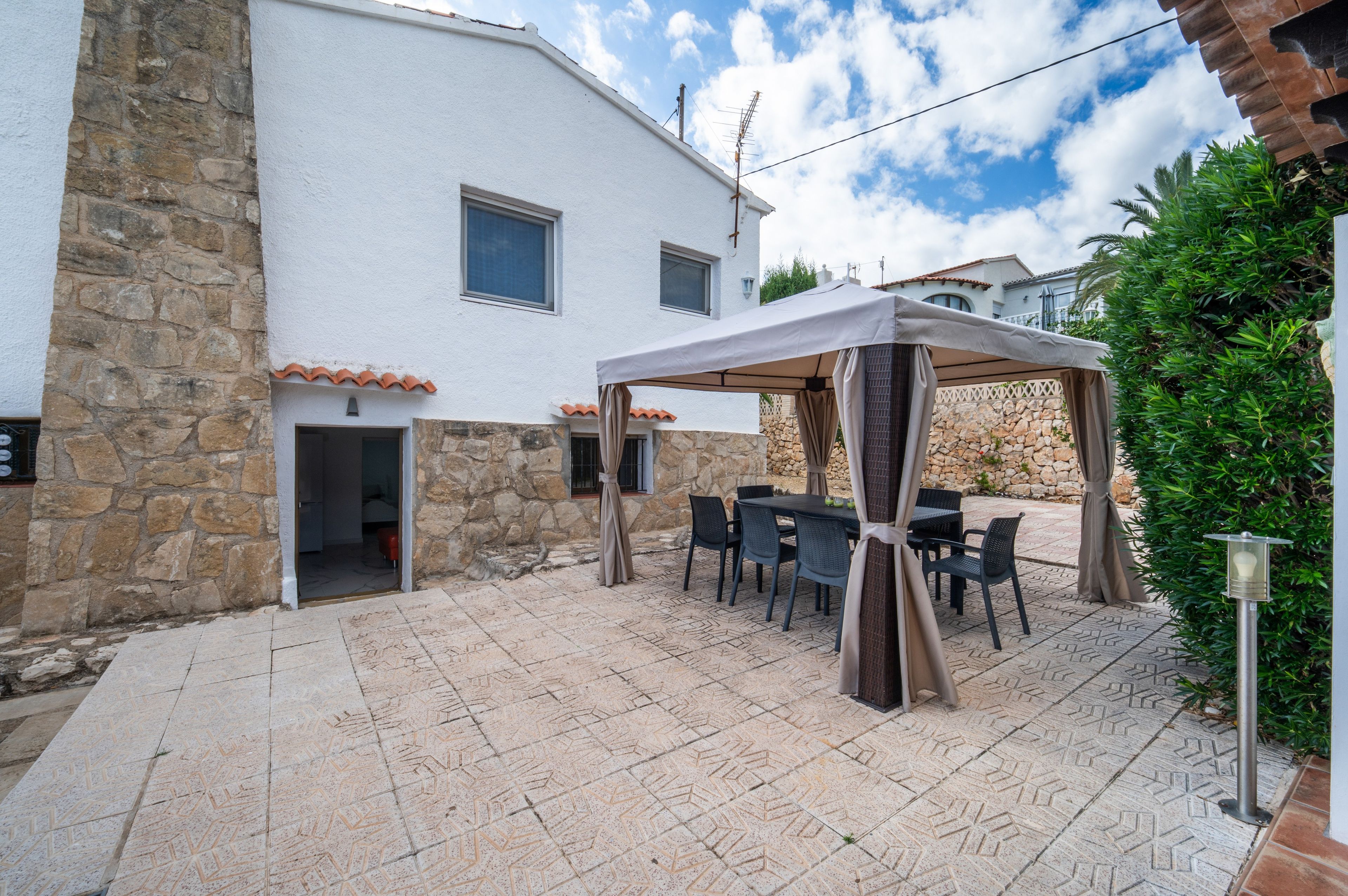 A covered outdoor seating area with tables and chairs, a stone wall, and a white building with windows.