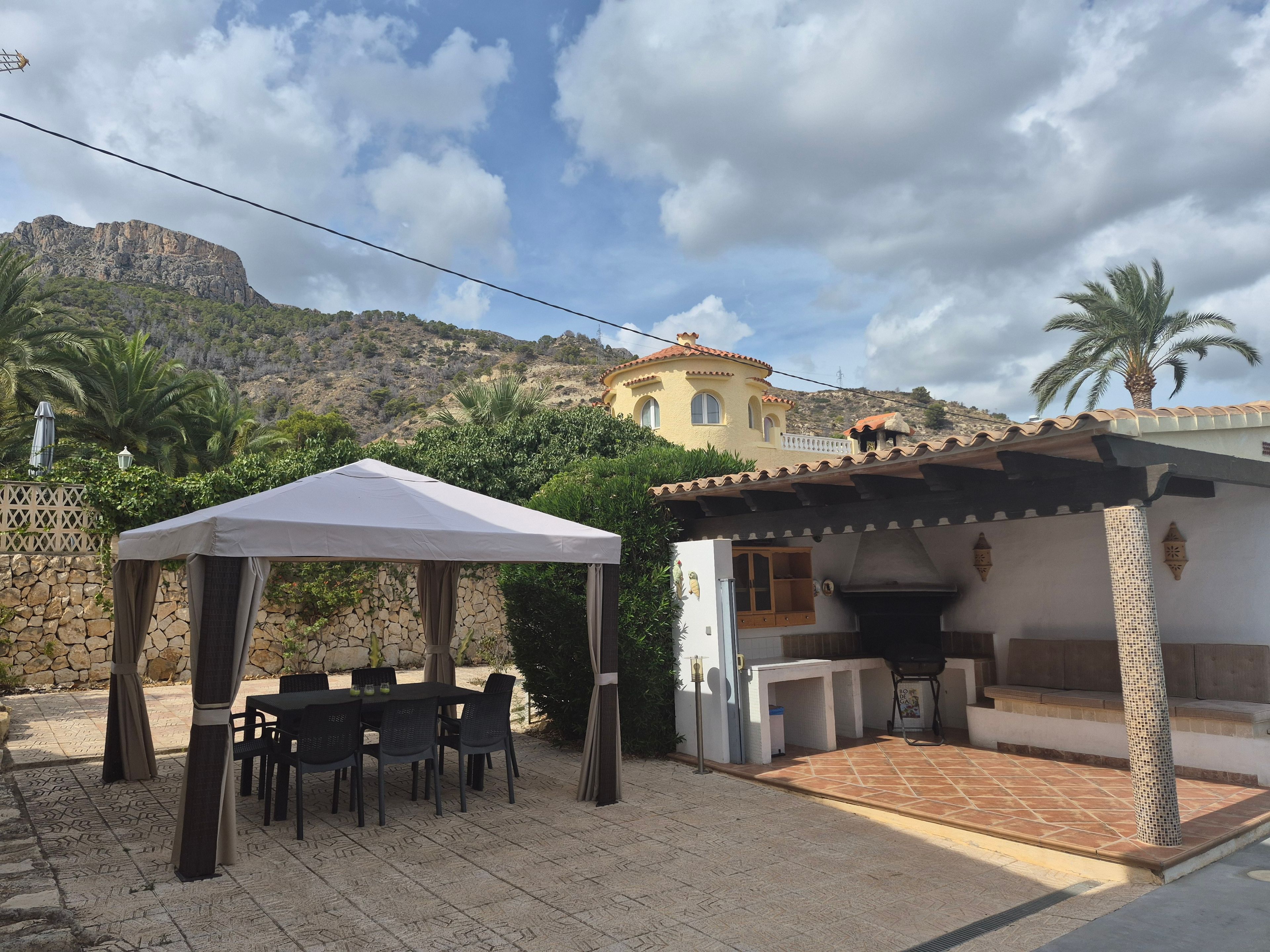 Un espacio patio al aire libre con una mesa y sillas, un área de estar cubierta y un edificio con techo de tejas.