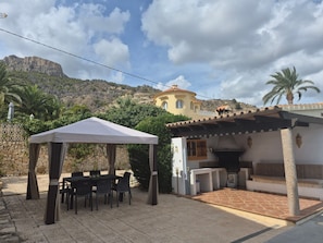 Un espacio patio al aire libre con una mesa y sillas, un área de estar cubierta y un edificio con techo de tejas.