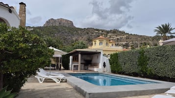 Zona de piscina con sillones, un área de estar techada y vistas a la montaña.
