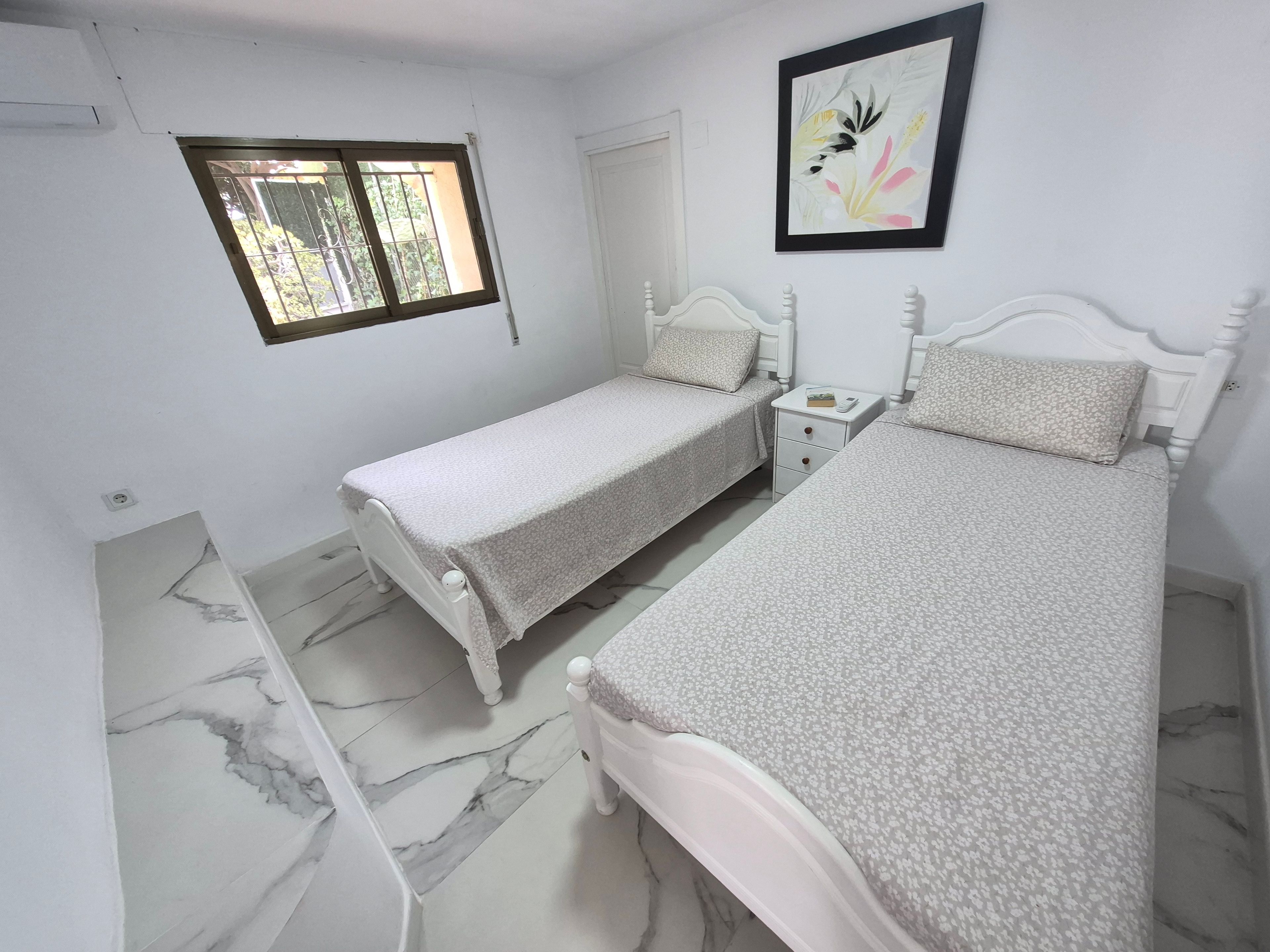 A bedroom with two single beds, a bedside table with a lamp, a window, and a framed picture on the wall.