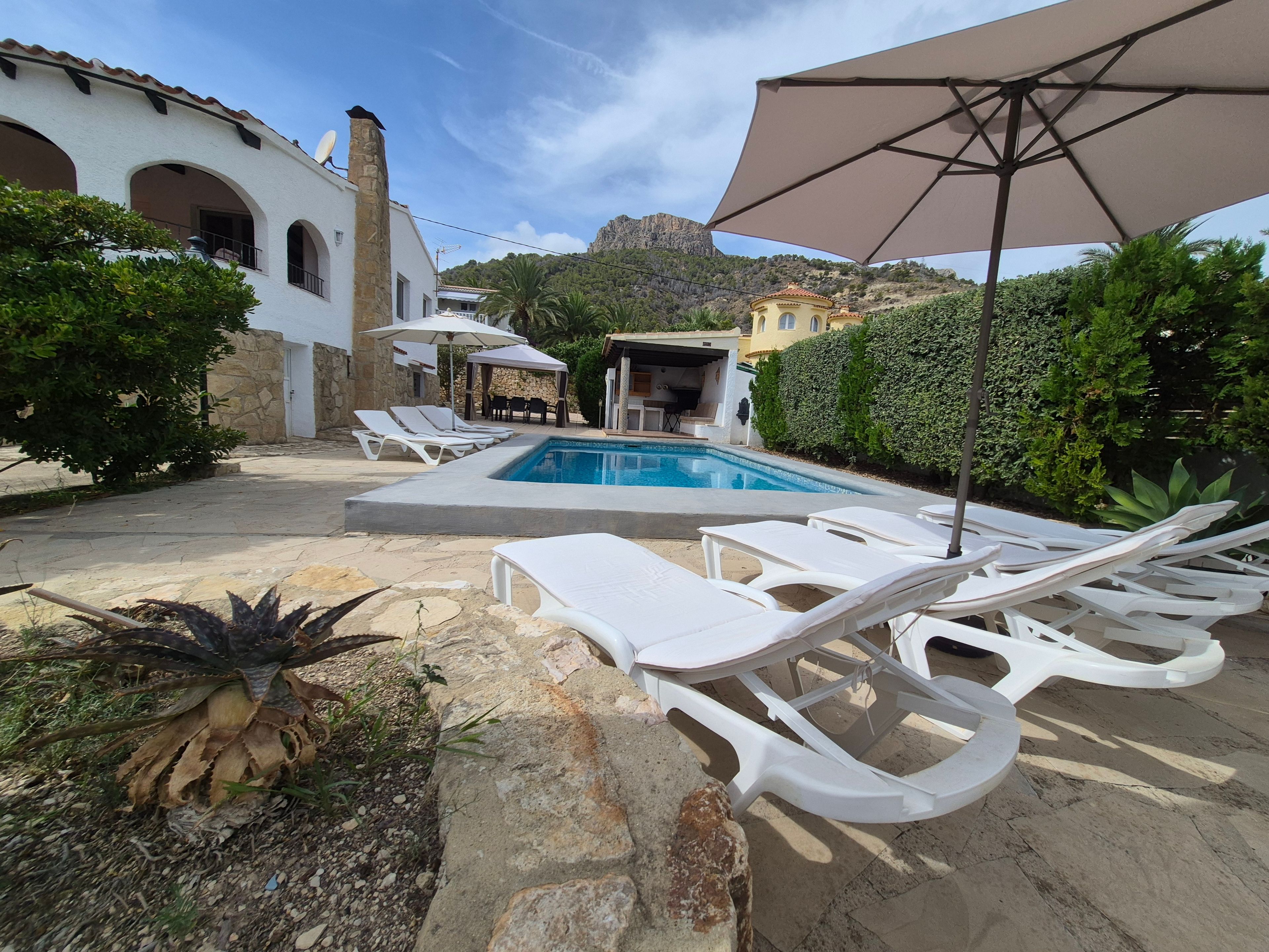 A pool area with white lounge chairs and a large umbrella, surrounded by greenery and a stone building.
