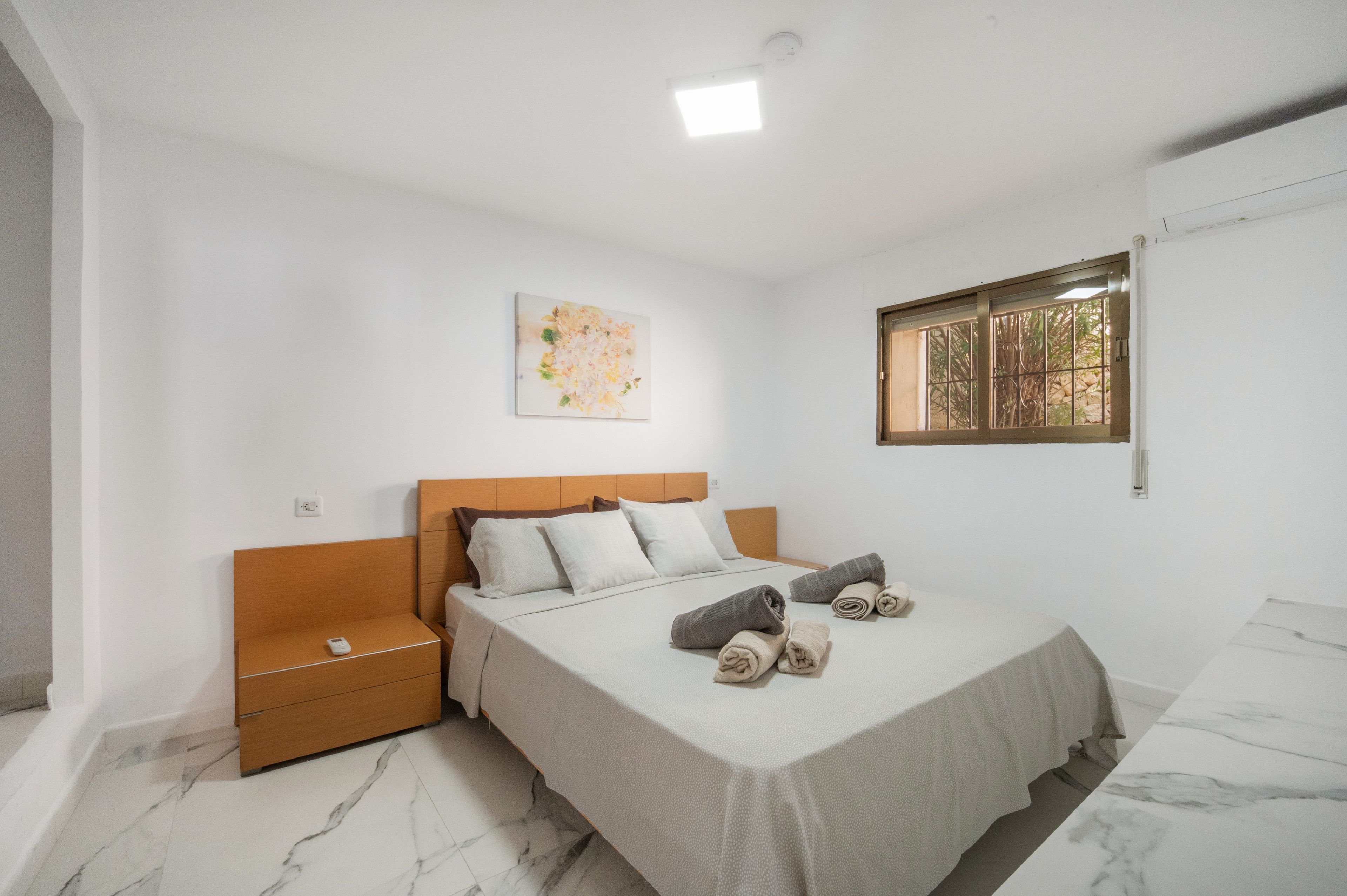 Een slaapkamer met een bed, een nachtkastje, een kunstwerk aan de muur, een raam met tralies en een marmeren vloer.