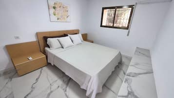 Une chambre à coucher avec un lit, des tables de chevet, un climatiseur fixé au mur, une fenêtre avec des barreaux et un tableau accroché au mur.