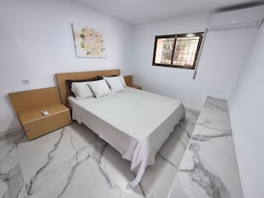 Un dormitorio con cama, mesitas de noche, aire acondicionado instalado en la pared, una ventana con rejas y un cuadro colgado.