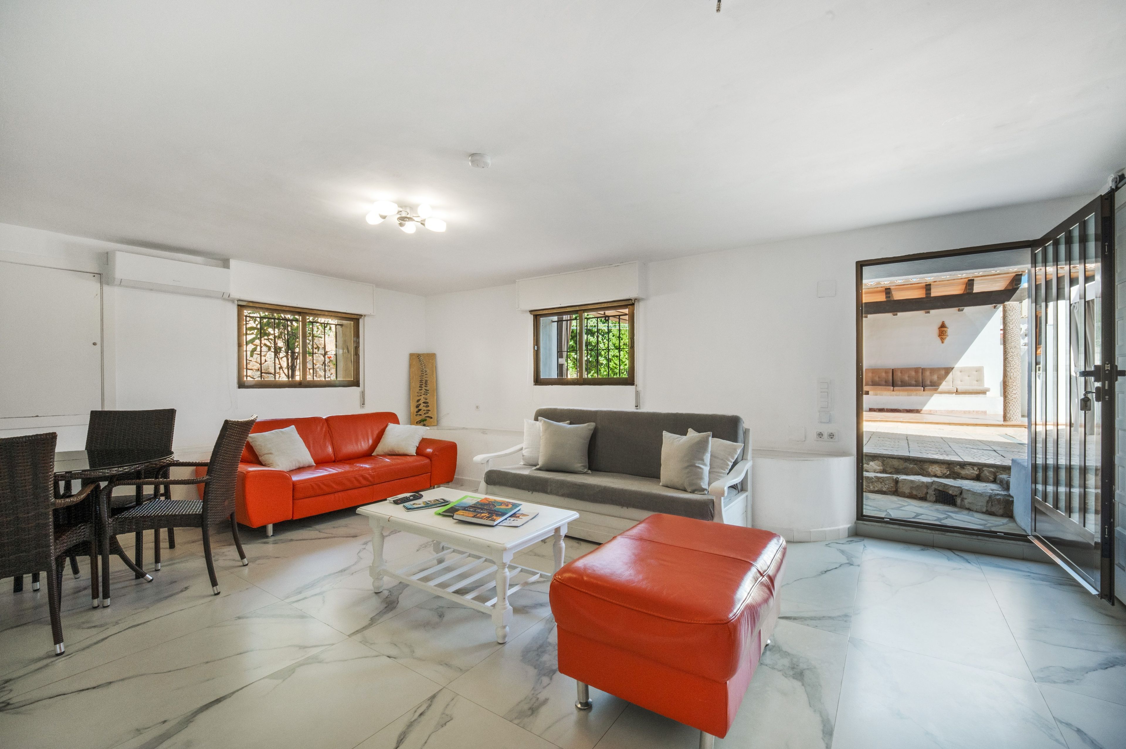 Ein Wohnzimmer mit einem roten Sofa, einem grauen Sofa, einem weißen Couchtisch und einem roten Hocker. Ein Fenster bietet Blick auf Grünflächen, und eine Tür führt nach draußen.