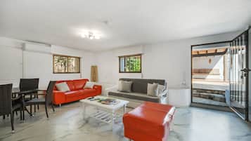 Ein Wohnzimmer mit einem roten Sofa, einem grauen Sofa, einem weißen Couchtisch und einem roten Hocker. Ein Fenster bietet Blick auf Grünflächen, und eine Tür führt nach draußen.