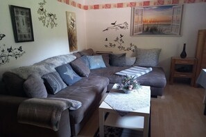A living room with a sectional sofa, a coffee table, and decorative wall art including a clock and a bird mural. - Haus Hoffmann Apartment A (Zell-Mosel)