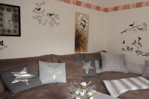 A living room with a brown sofa, star-shaped pillows, a clock, and wall art including birds and flowers. - Haus Hoffmann Apartment A (Zell-Mosel)