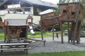 A playground with wooden play structures, including a slide and climbing elements, set against a building with a balcony and a sign that reads 'Cafe'. - Holiday apartment with 1 bedroom (Fischen)