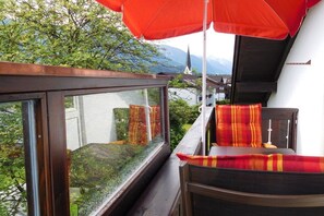 A balcony with a wooden railing, a red umbrella, and a view of a church and mountains. - Holiday apartment with 1 bedroom (Fischen)