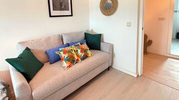 A living room with a beige sofa, green and blue pillows, a framed cityscape picture, and a decorative mirror on the wall.