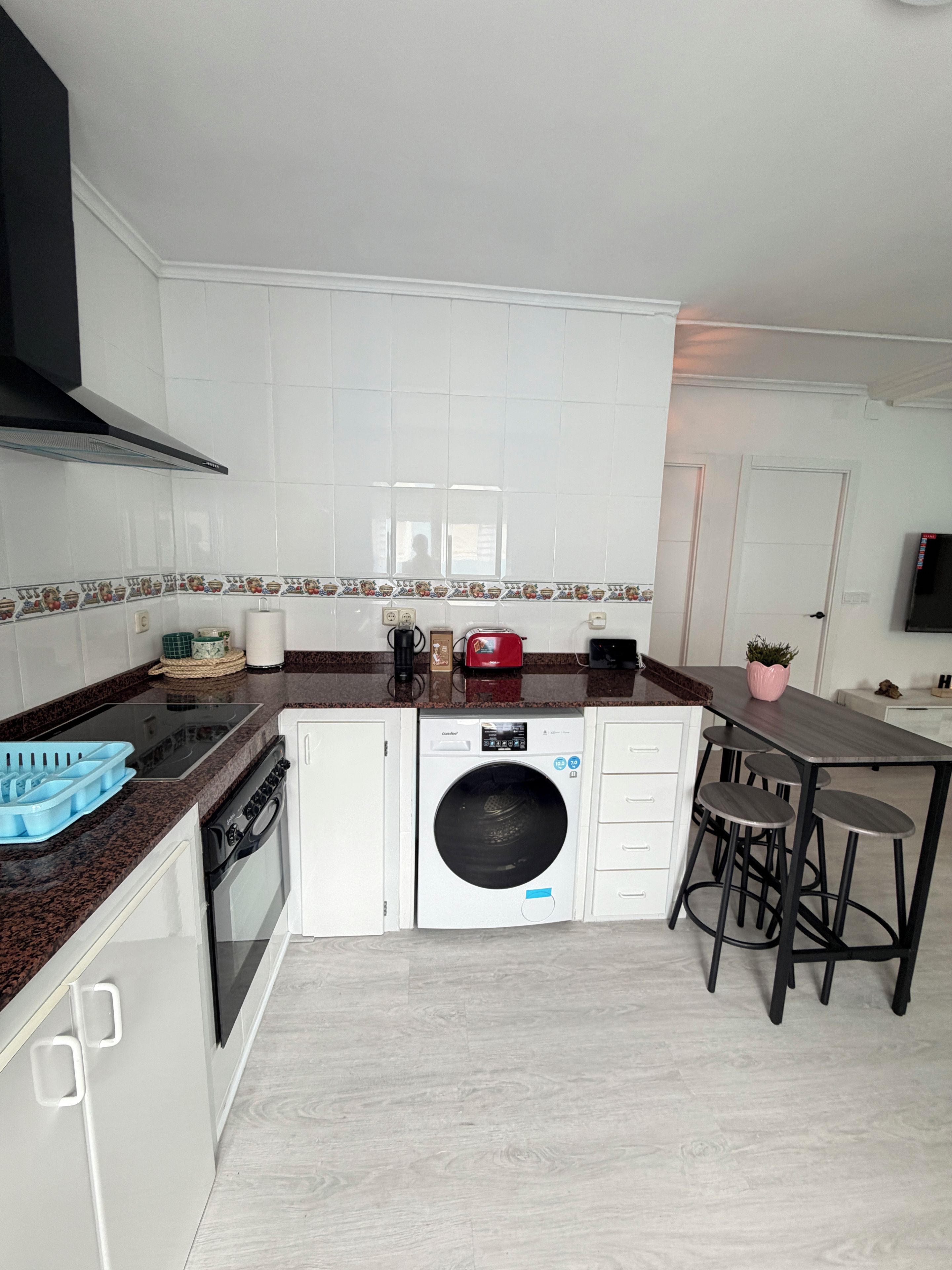A compact kitchen with white cabinets, a built-in washing machine, a stove, and a small dining area with stools.