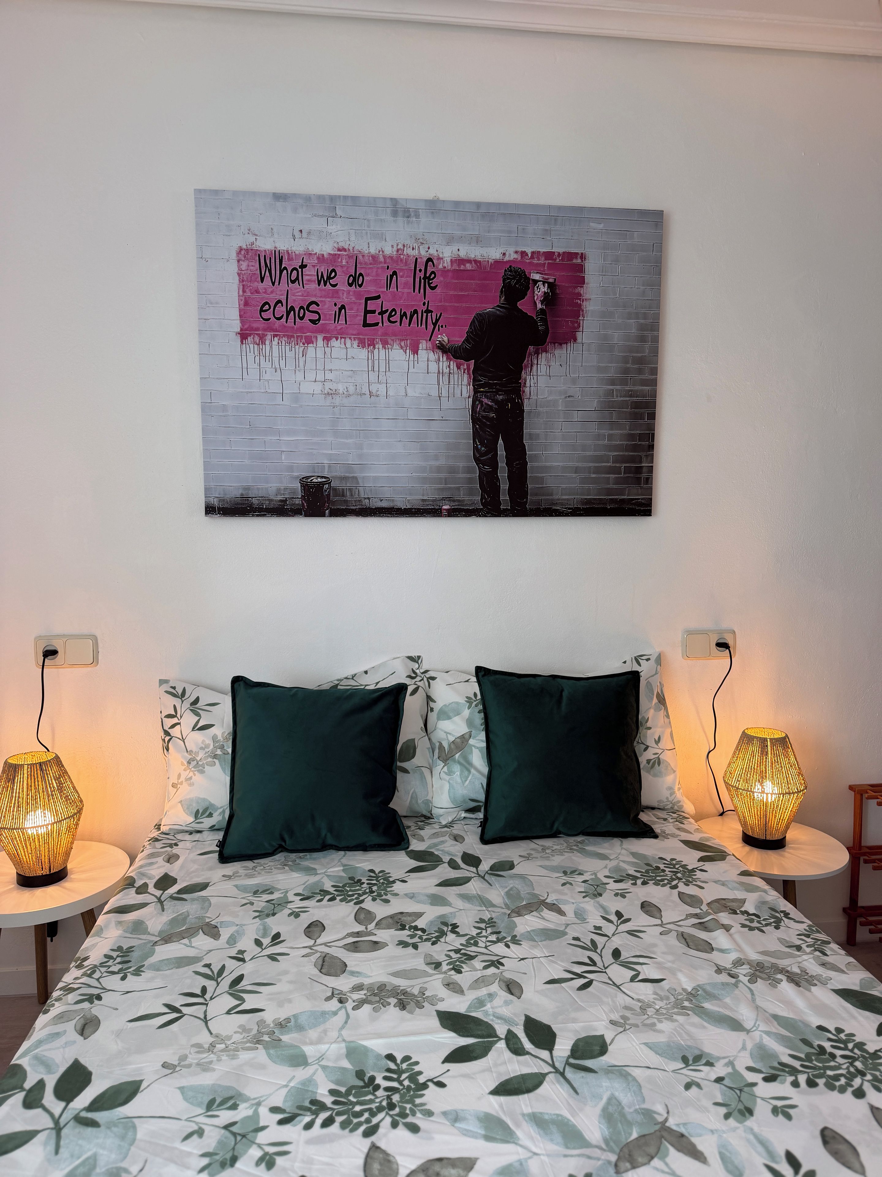 A bed with a floral bedspread, two green pillows, and a wall with a painting of a person painting a wall.