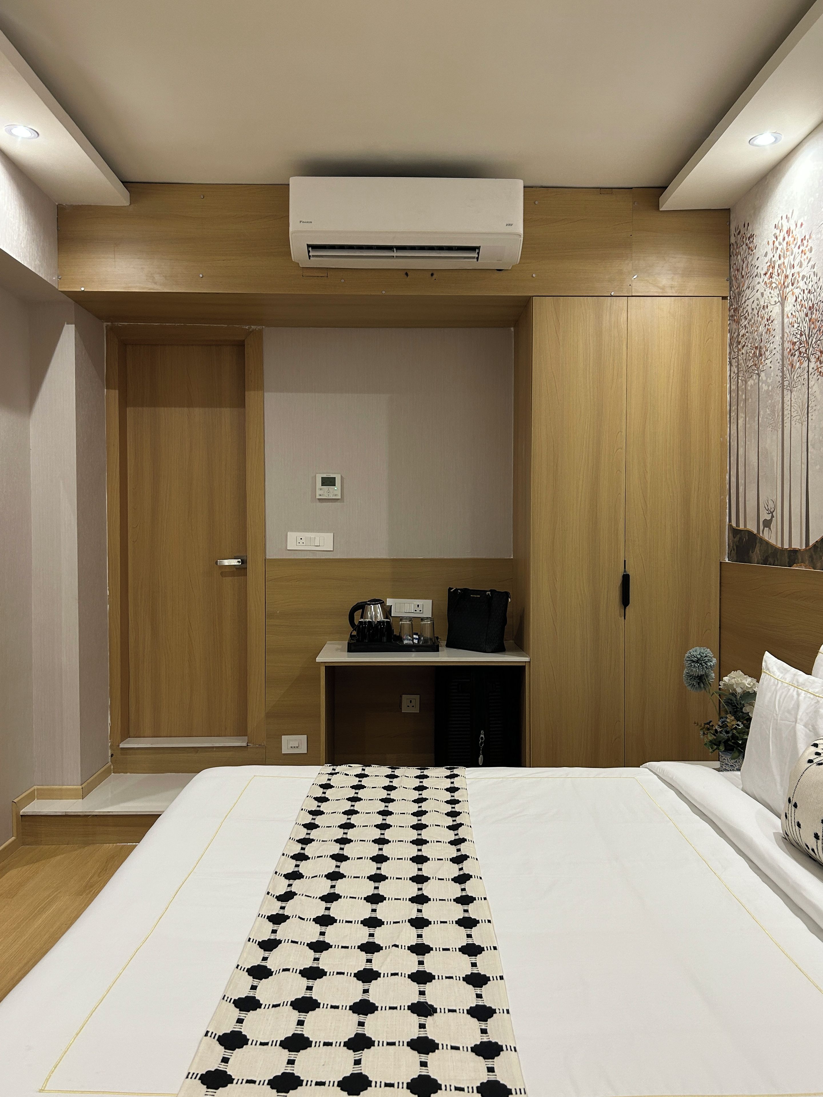 Habitación de hotel con una cama, una mesita de noche con tetera, un armario pequeño y un aire acondicionado de pared.