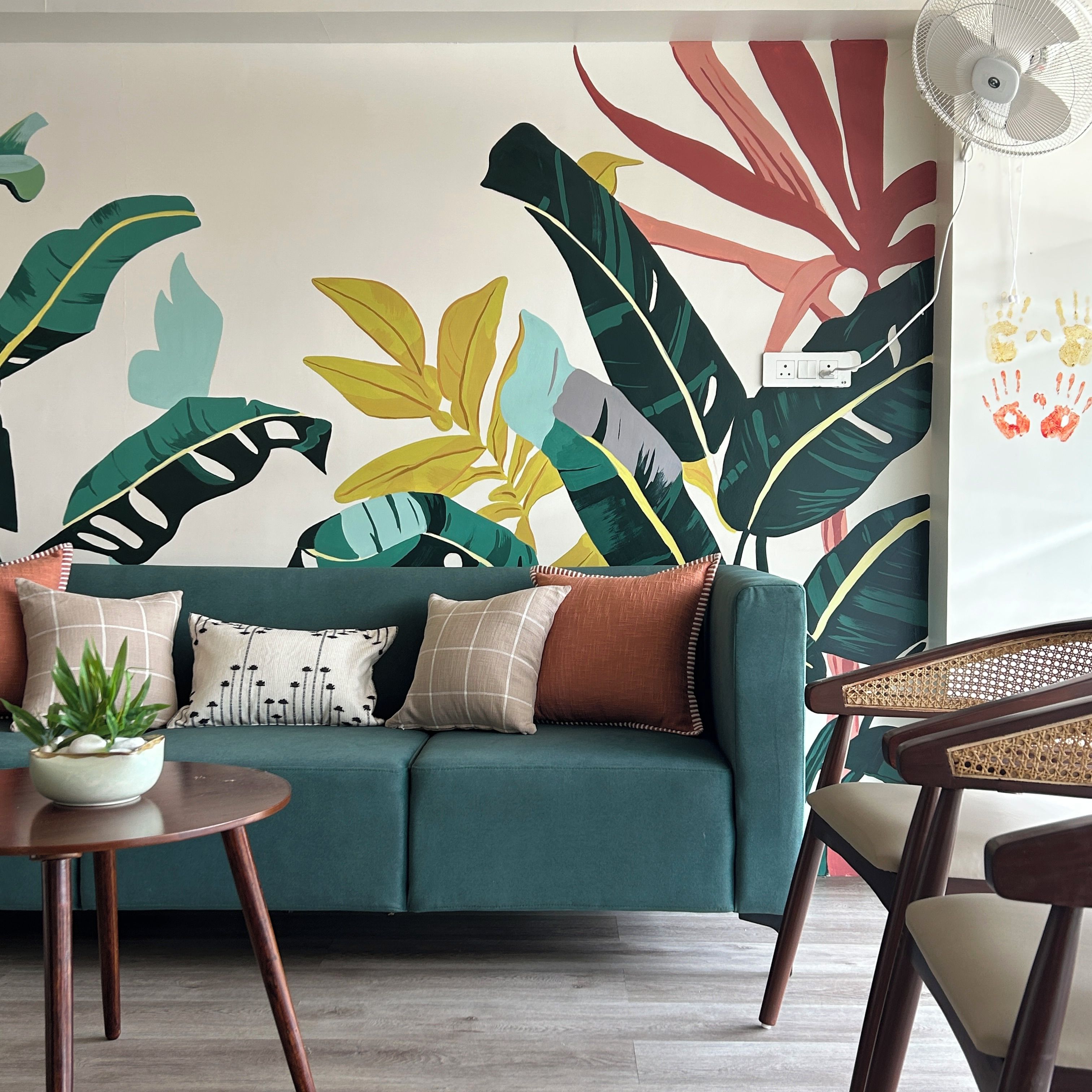 Una habitación con un sofá color turquesa, una mesa redonda de madera con una planta y una pared con un gran mural de hoja tropical.