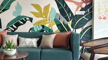 Una habitación con un sofá color turquesa, una mesa redonda de madera con una planta y una pared con un gran mural de hoja tropical.