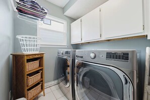 A laundry room with two front-loading washing machines, a wicker shelf with baskets, and a window with a view of the outside. - Miramar Beach | Beach Access & Fire Pit (Miramar Beach)